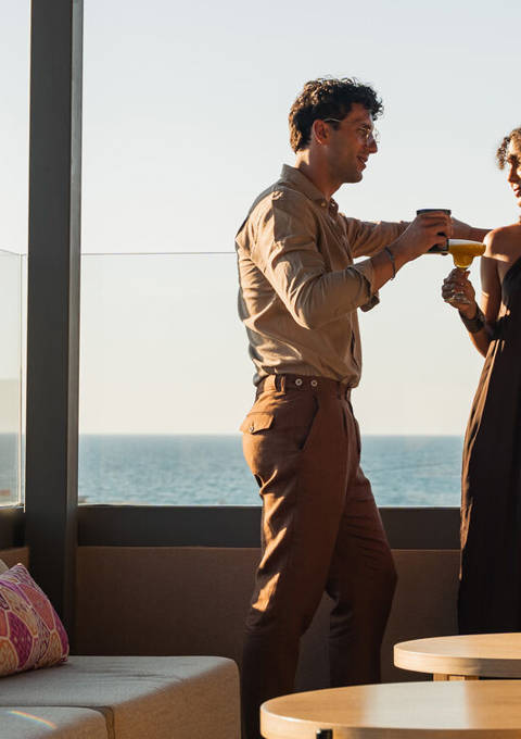 Couple toasting with cocktails at the rooftop lounge with the sea behind them