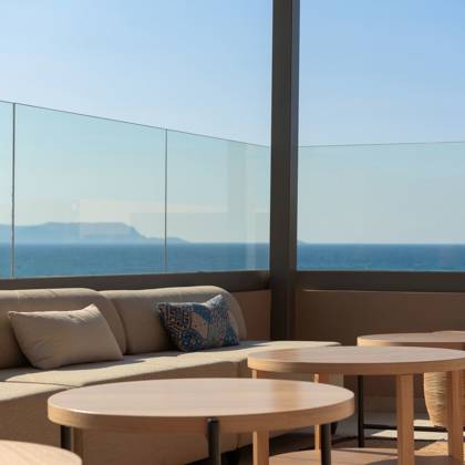 Canapé lounge avec coussins et tables basses en bois derrière une balustrade vitrée face à la mer