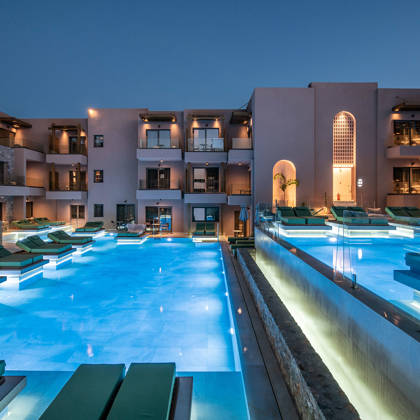 Piscines extérieures illuminées entourées de lits de repos et transats, avec les bâtiments de l'hôtel au crépuscule