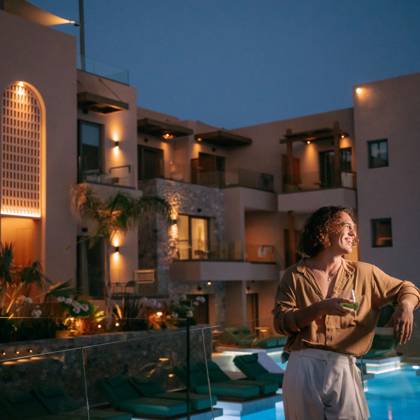 Homme souriant, un verre à la main, près de la piscine illuminée et des balcons éclairés de l'hôtel