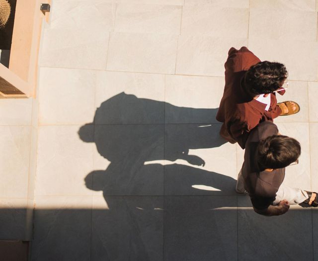 Two guests walking across a sunlit terrace at Paralos Venus Suites, casting long afternoon shadows.